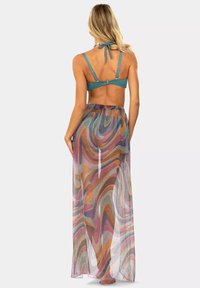 Kobieta w turkusowym biustonoszu bikini oraz długiej, przezroczystej spódnicy w multikolorowy wirujący wzór, stoi boso z plecami zwróconymi do przodu.