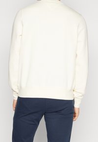 Sudadera color crema con un cuello redondo clásico, puños y dobladillo acanalados, que presenta una textura suave y un corte relajado, usada con pantalones de color marino.