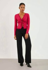 Cardigã longo vermelho de mangas largas com bainha rendilhada e laço frontal, combinado com calças pretas de perna larga e sapatos de salto pretos de bico fino.