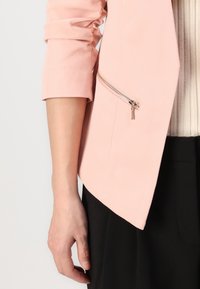 Lichtroze blazer met driekwart mouwen, voorzien van een schuin afgeronde zoom en een zilveren ritssluiting detail op de zak. Gecombineerd met een geribd topje en zwarte broek.