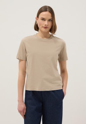 Mujer con cabello corto y castaño, lleva una camiseta beige de manga corta y pantalones azul oscuro, de pie contra un fondo liso y claro.