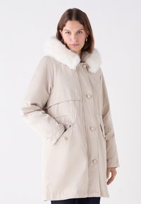 Beige parka met een nepbontkraag, voorzien van een knoopsluiting aan de voorkant, zijzakken en een ontspannen pasvorm. Soepele textuur, lange mouwen en een minimalistische stijl.