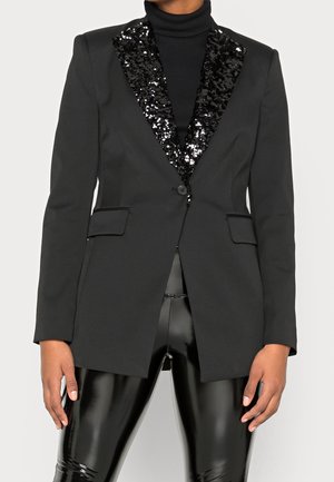 Schwarzer Blazer mit paillettenbesetzten Revers, getragen über einem schwarzen Rollkragenpullover und glänzenden schwarzen Vinylhosen, Hände locker an den Seiten.