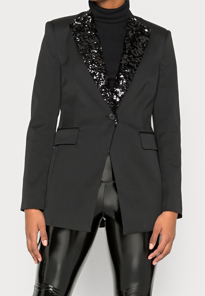 Blazer noir avec revers à sequins porté sur un col roulé noir et un pantalon en vinyle noir brillant, mains détendues sur les côtés.