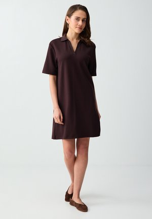 Donna in piedi che indossa un vestito polo marrone scuro fino al ginocchio con maniche corte e scarpe marroni basse su uno sfondo chiaro semplice.