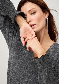 Glitrende sort bluse med lange, puffede ærmer, scalloped manchetter og dyb V-udskæring. Har en metallisk tekstur og glat design.