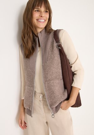 Brown fleece vest with knitted shoulders and zipper, paired with a beige top and light-colored pants; holding a dark brown bag.