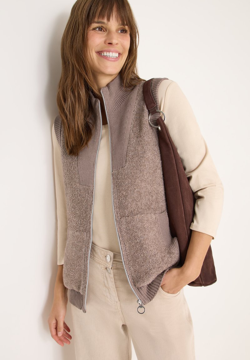 Braune Fleece-Weste mit gestrickten Schultern und Reißverschluss, kombiniert mit einem beigen Oberteil und hellen Hosen; hält eine dunkelbraune Tasche.