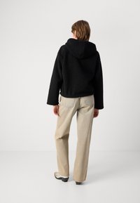 Casaco com capuz forrado a fleece preto, de corte relaxado, combinado com calças bege de perna reta e calçado preto de bico quadrado.