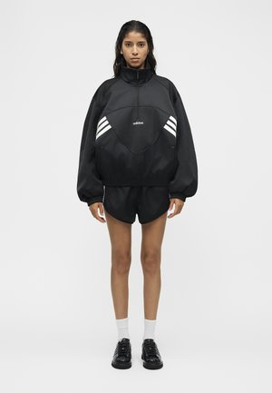 Vrouw staat frontaal gekleed in een zwarte Adidas trui met witte strepen, zwarte korte broek, witte sokken en zwarte schoenen tegen een effen achtergrond.