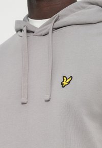 Jasnoszara bluza z kapturem ze sznurkiem, z małym żółtym haftowanym logo ptaka na lewej piersi. Miękka, teksturowana tkanina.