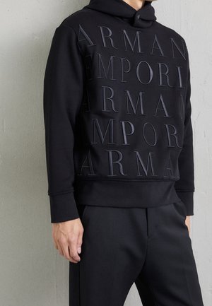 Sweat à capuche noir avec grand texte embossé ARMANI EMPORIO porté par une personne debout contre un mur gris, associé à un pantalon noir.