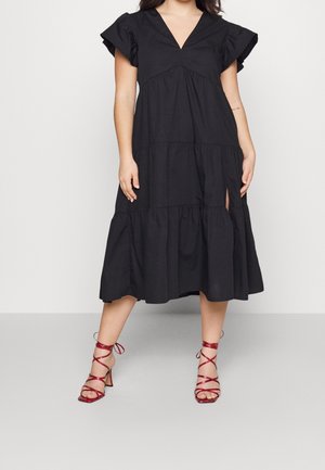 Kvinde iført en sort tiered midi-kjole med flagrende ærmer og en frontslids, kombineret med røde, snørede højhælede sandaler.