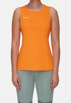 Femme portant un haut sans manches orange vif avec le logo blanc "Mammut" et un pantalon ajusté gris clair, debout devant un fond uni.