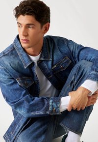 Denim jacket in medium blue with a classic fit, button front, dual chest pockets, and subtle fading. Worn over a white t-shirt.