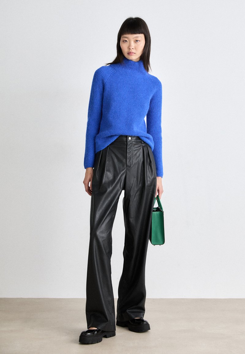 Pull en maille côtelée bleu avec col haut, associé à un pantalon large en cuir noir. Tient un petit sac à main vert. Chaussures noires épaisses.