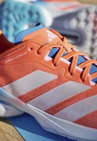 Primo piano di una scarpa da corsa Adidas arancione e bianca con fodera interna blu e lacci su una superficie in legno chiaro.