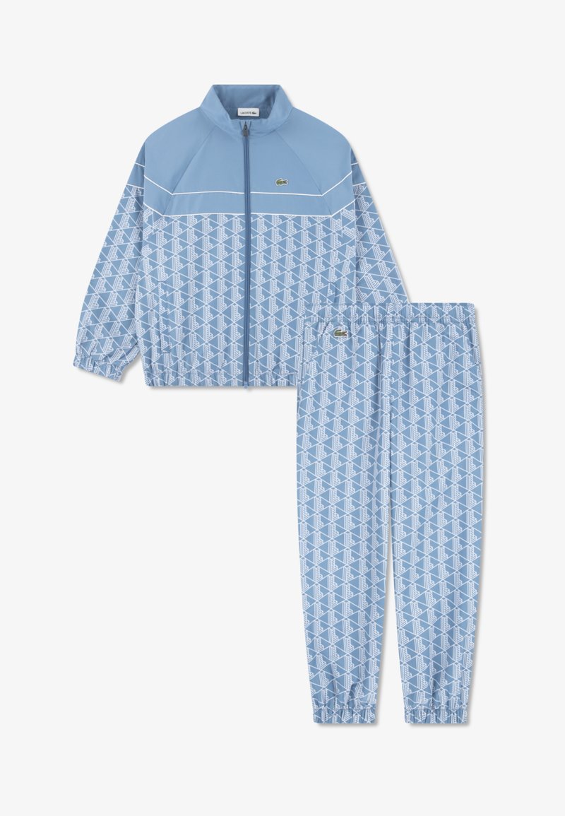 Survêtement bleu clair en tissu lisse, avec un motif géométrique blanc. La veste présente un col montant, une fermeture éclair et des poignets élastiques.