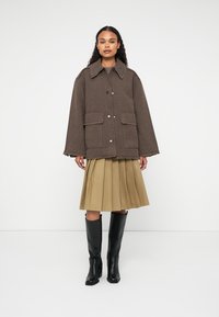 Vrouw in een bruin oversized jack, beige geplooide rok en zwartekante laarzen tot de knie, staande tegen een effen witte achtergrond.