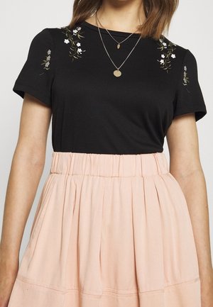 Czarna koszula z krótkim rękawem z kwiatowym haftem na ramionach, połączona z jasnoróżową spódnicą z marszczonym paskiem w talii i wielowarstwowym fasonem.