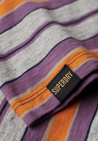 Stribet stof i grå, lilla, orange og marineblå. Har en foldet kant med en sort etiket, der st�år "SUPERDRY" i orange tekst. Blød tekstur.