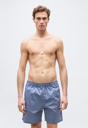 Joven con cabello rizado, sin camisa, vistiendo pantalones cortos de baño azul claro, de pie mirando hacia adelante contra un fondo claro y uniforme.