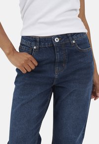 Granatowe dżinsy o widocznych szwach, pięć kieszeni, z zapięciem na guzik, pokazane z ręką umieszczoną na biodrze.