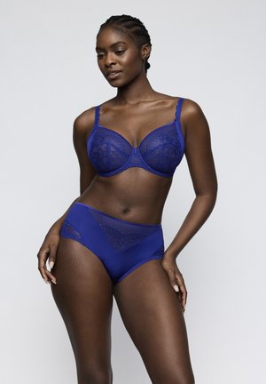 Donna con capelli intrecciati che indossa un reggiseno di pizzo blu abbinato e slip a vita alta, in piedi contro uno sfondo chiaro e uniforme.