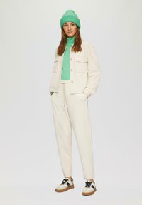 Frau trägt eine leuchtend grüne Mütze und einen Rollkragenpullover unter einer cremefarbenen Jacke, passenden cremefarbenen Hosen und weißen Sneakern mit schwarzen Details.