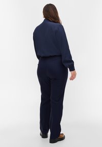 Kvinna med långt brunt hår som vänder ryggen mot kameran, klädd i en marinblå blus, matchande byxor och bruna skor mot en enkel vit bakgrund.