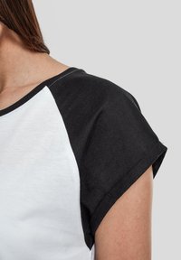 Camiseta de manga corta en blanco y negro con cuello redondo, que presenta mangas raglán negras y costuras en contraste a lo largo del cuello.