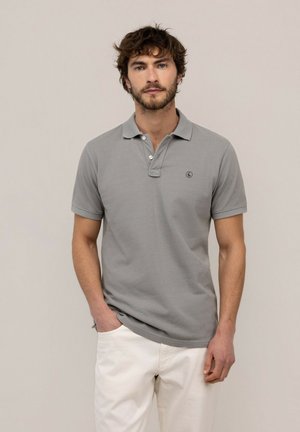 Hombre con cabello rizado y barba que lleva una camisa polo gris de manga corta y pantalones blancos, de pie con una mano en el bolsillo.