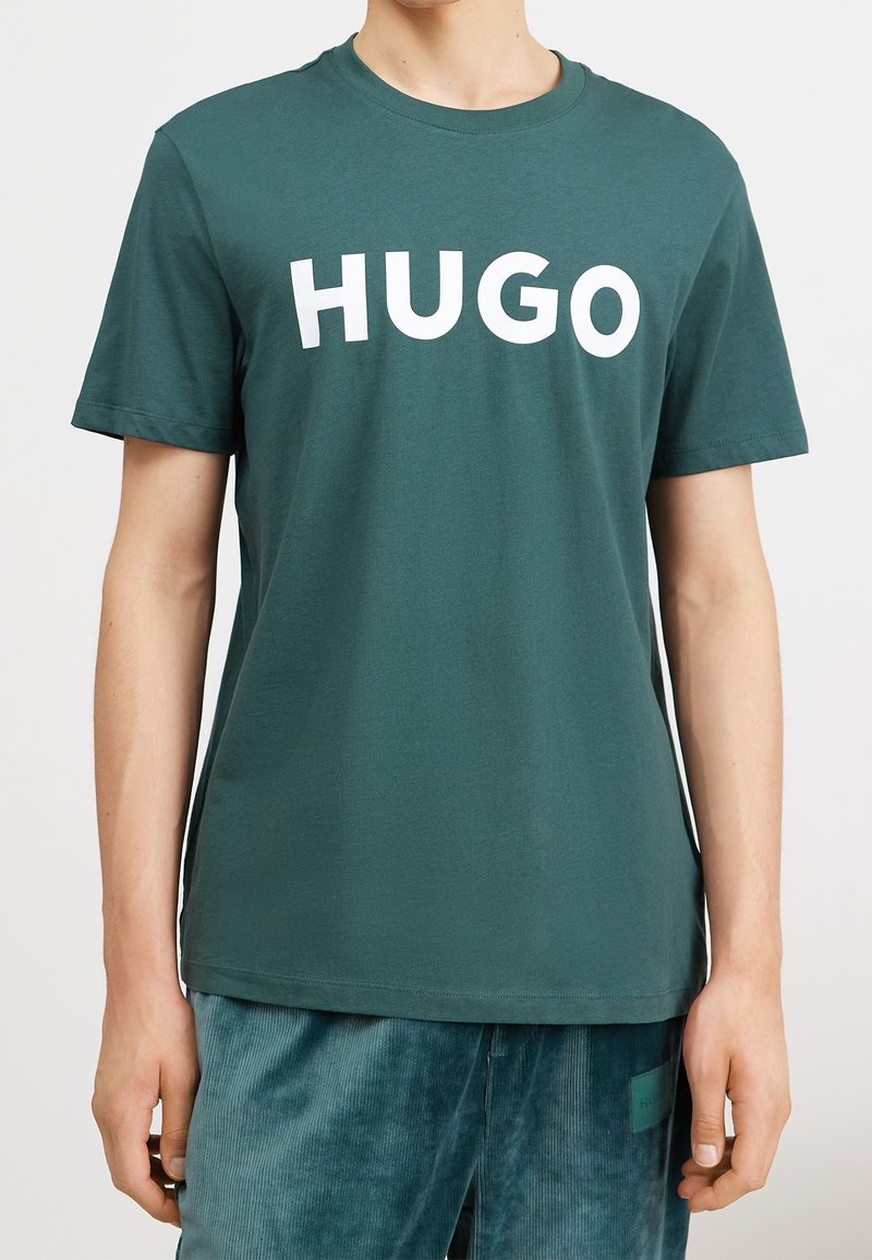 T-shirt en coton vert avec un grand texte blanc "HUGO" sur la poitrine. Manches courtes et encolure ras du cou. Texture lisse.