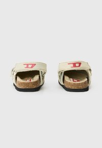 Gai bēšas slip-on sandalītes ar sarkaniem akcentiem, korķa zolīte, tekstūras siksnas un melna gumijas zole. Aizmugures skats, kas parāda dizaina detaļas.