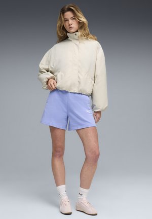 Kobieta ubrana w beżową, pikowaną kurtkę, jasnofioletowe spodenki, białe skarpetki Puma i jasnoróżowe trampki stoi na tle szarego gradientu.