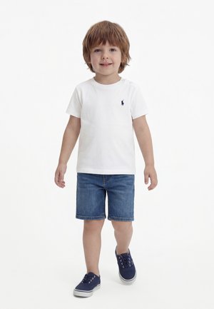 Jeune garçon aux cheveux châtain clair portant une chemise blanche à manches courtes, un short en jean et des baskets bleu marine, marchant vers l'avant sur un fond blanc.