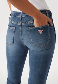 Jeans skinny azul oscuro con una cintura alta, costuras visibles y dos bolsillos traseros. Un pequeño logo triangular rojo está en el bolsillo derecho.