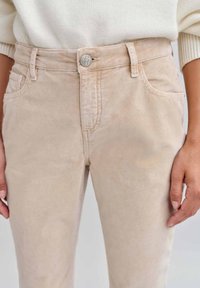 Beige Cord-Hosen mit einer glatten Textur, ausgestattet mit einem vorderen Knopfverschluss, Gürtelschlaufen und seitlichen Taschen.