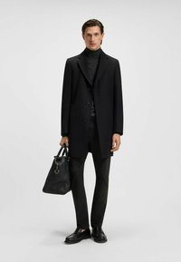 Czarny wełniany płaszcz o dopasowanym kroju, z trzema guzikami i klapami. W zestawieniu z czarnym golfem, dopasowanymi spodniami i skórzanymi butami, trzyma torbę.