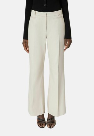 Vrouw die crèmekleurige, wijduitlopende broek met hoge taille draagt, een zwart shirt met lange mouwen en zwarte open schoenen met studs, tegen een effen achtergrond.