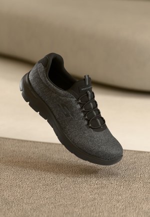 Mørkegrå Skechers slip-on sneaker med sort sål og elastiske snørebånd, der svæver over beige tekstureret overflade og baggrund.