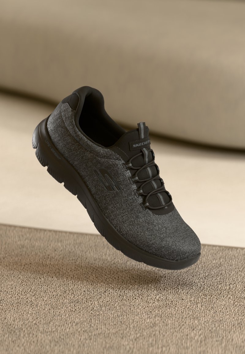 Basket Skechers slip-on gris foncé avec semelle noire et lacets élastiques flottant au-dessus d'une surface texturée beige et d'un arrière-plan.