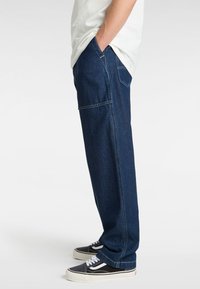 Pantalones de mezclilla azul oscuro con corte relajado, que cuentan con bolsillos laterales y costuras en contraste. Combinados con zapatillas negras y una camiseta blanca.