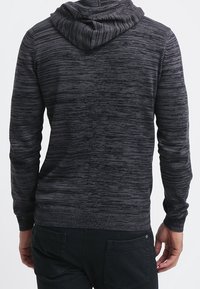 Šedá pletená mikina s texturovanými vodorovnými pruhy; má kapuci se stahovací šňůrkou a žebrované manžety a lem. Natažena s tmavými kalhotami.