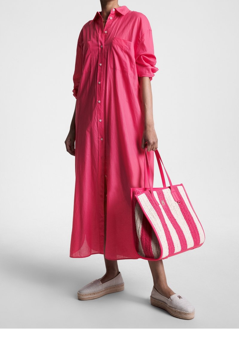 Personne portant une longue robe rose boutonnée, tenant un grand sac fourre-tout rose et blanc rayé, assortie à des espadrilles de couleur claire.