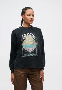 Czarna bluza z teksturowanej tkaniny, z kolorowym wzorem graficznym przedstawiającym słowo "ROXY" oraz motyw oceanu. Do tego brązowe spodnie.