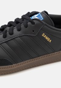 Schwarzer Ledersneaker mit drei schwarzen Streifen, goldenem "SAMBA"-Schriftzug und brauner, strukturierten Gummisohle. Verfügt über runde schwarze Schnürsenkel und ein blaues Logoband.