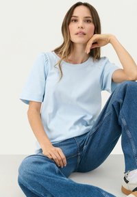 Top corto de manga corta azul claro con escote redondo y hombros ligeramente abullonados, combinado con jeans azules de cintura alta, sentado frente a un fondo neutro.