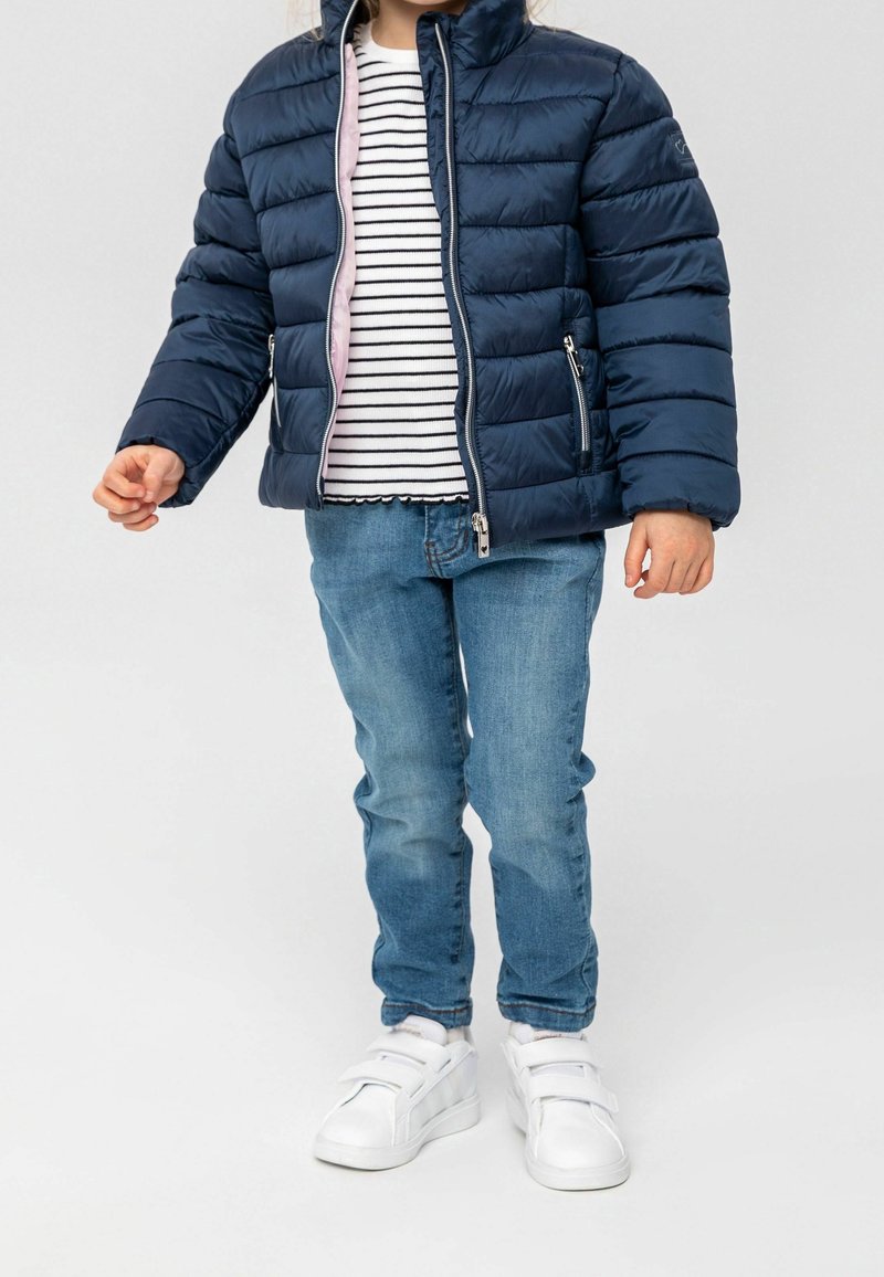 Enfant portant une veste matelassée bleu marine par-dessus une chemise rayée blanche et noire, un jean bleu, et des baskets blanches à scratchs sur un fond uni.