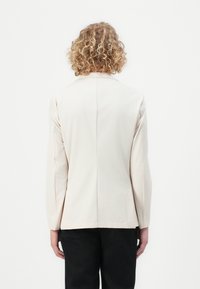 Béžový přizpůsobený blazer s hladkou texturou, dlouhými rukávy a rovnými liniemi, se středovým švem a dvěma bočními kapsami.